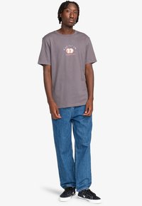 Camiseta de algodón gris con un estampado gráfico que presenta ojos y texto, combinada con jeans de mezclilla azules holgados y zapatillas negras con suelas blancas.