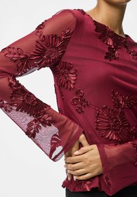 Burgunderfarget bluse med gjennomsiktige, broderte ermer med blomsterstiler og klokkeermer. Jevn tekstur med intrikate detaljer over hele.