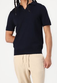 Granatowa koszulka polo z krótkim rękawem, dekoltem w serek, prążkowanym kołnierzykiem i dołem, wykonana z gładkiego materiału, zestawiona z beżowymi spodniami ze sznurkiem.