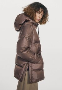 Kvinde med krøllet hår iført en brun polstret hættetrøje og beige nederdel, stående med hænderne i jakkelommerne, kiggende sidelæns.