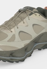Chaussure de sport beige et grise avec panneaux en mesh, semelle texturée unique et système de laçage sécurisé de type bungee.