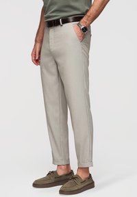 Homme portant un pantalon beige clair retroussé, des chaussures bateau vert olive, une ceinture foncée, une montre-bracelet, et un t-shirt vert, debout avec une main dans la poche.