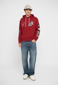 Rød hættetrøje med logo og lapper, snoretræk i hætten, front-kangaroo lomme. Sammen med lyseblå jeans og hvide sneakers.