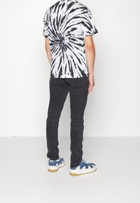 Osoba stojí s zády k fotoaparátu, oblečena v tričku s černobílým tie-dye vzorem, tmavých džínách a modro-bílých teniskách.