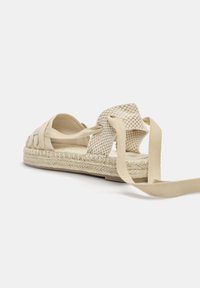 Béžová espadrilová sandál s tkaným designem popruhů, texturovanou tkaninou, plochou jutu podrážkou a dlouhými plátěnými šňůrkami na zadní straně pro bezpečné uchycení.