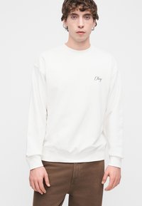 Λευκό φούτερ με ριμπ μανσέτες και τελείωμα, διαθέτει διακριτική κεντημένη επιγραφή "Obey" στο στήθος, συνδυασμένο με καφέ παντελόνι.