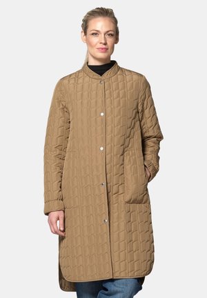 Woman wearing a long tan quilted coat with snap buttons and side pockets, standing with one hand in pocket against a plain background.