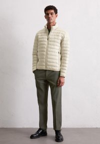 Jasnobeżowa pikowana kurtka z wysokim kołnierzem, zamkiem błyskawicznym i bocznymi kieszeniami, w zestawieniu z oliwkowozielonymi spodniami i czarnymi sznurowanymi butami.