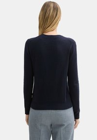 Marineblauer Pullover mit langen Ärmeln, gerippten Bündchen und Saum, strukturiertem Stoff und einem Rundhals. Tragen über grauen schmal geschnittenen Hosen.