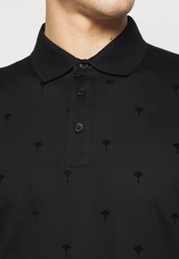 Polo noir en tissu doux présentant un motif texturé de petits motifs en forme de feuilles. Patte de boutonnage à trois boutons avec des boutons assortis.