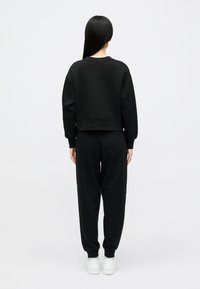 Άτομο με μακριά μαύρα μαλλιά στέκεται με το βλέμμα στραμμένο προς τα πίσω, φορώντας μια μαύρη φούτερ, μαύρα παντελόνια φόρμας και λευκά αθλητικά παπούτσια σε ένα απλό φόντο.