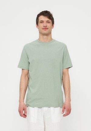 Mann som står og har på seg en lysegrønn t-skjorte med korte ermer og hvite bukser mot en ensfarget hvit bakgrunn.