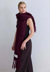 Bordeaux wollen sjaal met franjes aan de uiteinden, voorzien van een label op witte stof. Losjes gedragen over een donkere top, tegen een neutrale achtergrond.