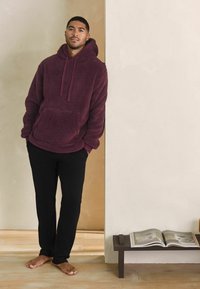 Bordeaux fleece hoodie met kangoeroezak; gecombineerd met zwarte broek. Blote voeten, leunend tegen een lichtgekleurde muur.