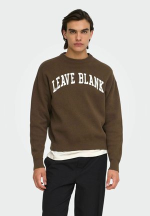 Ung mand iført en brun sweater med teksten "LEAVE BLANK", hvid undertrøje og sorte bukser, stående mod en ensartet baggrund.