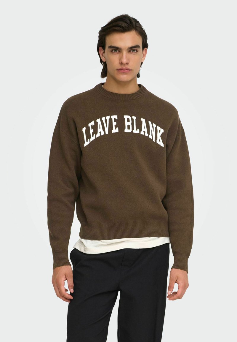 Jeune homme portant un pull marron avec le texte "LEAVE BLANK", un sous-vêtement blanc et un pantalon noir, debout devant un fond uni.