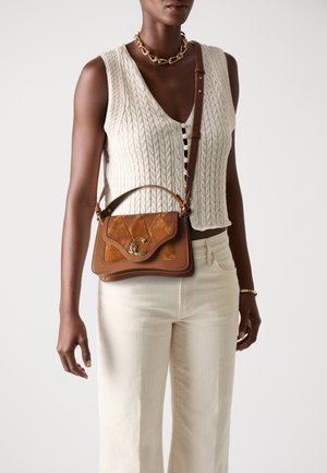 Tan leather handbag with a quilted texture, gold hardware, and a curved flap closure. Worn crossbody over a knitted cream top and beige pants.