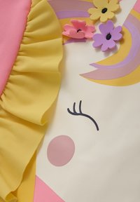 Le produit présente un tissu rose avec des volants jaunes. Il comprend des embellissements floraux colorés et un design facial minimaliste sur fond crème.