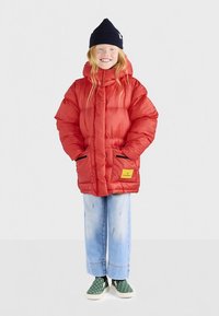 Dijete nosi crvenu puffersku jaknu, svijetloplave traperice, zelene cipele bez vezica s bijelim točkicama i crnu kapu, stoji i smiješi se.