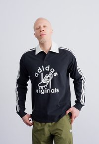 Cămașă polo neagră cu mâneci lungi, cu dungi albe pe mâneci, având un logo alb adidas și un design grafic în față; asortată cu pantaloni verzi.