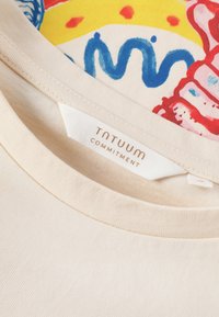 Nahaufnahme des Ausschnitts eines beigen T-Shirts, das ein „TNTUUM COMMITMENT“-Label und darunter einen farbenfrohen Stoff mit blauen, roten und gelben abstrakten Mustern zeigt.