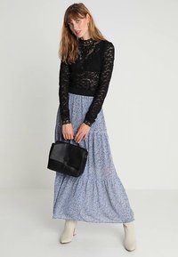 Fekete csipkés, hosszú ujjú felsőt, kék virágmintás maxi szoknyát, fehér csizmát viselő nő, aki egy fekete táskával áll.