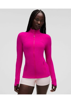 Donna con lunghe trecce che indossa una giacca aderente rosa acceso con zip e pantaloncini rosa chiaro, in piedi davanti a uno sfondo chiaro e uniforme.
