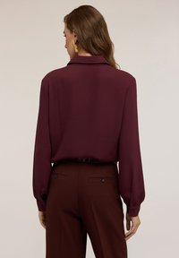 Blusa a maniche lunghe in prugna con colletto, tessuto morbido e polsini con bottoni, abbinata a pantaloni coordinati con tasche posteriori e bottoni.