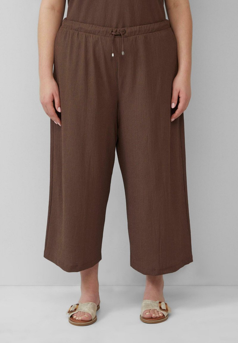 Bruine, wijde broek van textuurstof met een elastische tailleband en trekkoorden. Gecombineerd met lichtgekleurde slidesandalen.