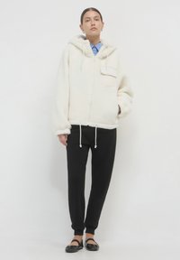 Felpa con zip colore crema, dotata di tasca frontale, abbinata a pantaloni jogger neri e scarpe nere piatte. Vestibilità ampia e texture morbida.