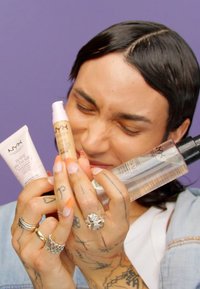 Tre NYX-kosmetiska produkter: en rosa kräm i tub, ett brunt serum med pump och en klar sprayflaska. Alla produkter har varumärkesetiketter.