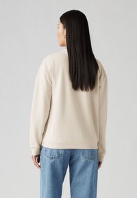 Felpa beige con polsini e bordo a coste, realizzata in un morbido tessuto. Abbinata a jeans a vita alta di colore blu chiaro, che mostrano un fit rilassato.