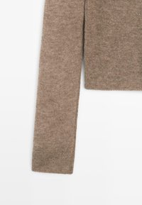 Langer Ärmel eines weichen, gestrickten beigen Pullovers mit einem gerippten Bündchen, gezeigt vor weißem Hintergrund.