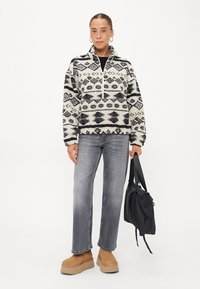 Pull en polaire ivory avec un motif géométrique noir, col demi-zip, associé à un jean gris et des bottes à plateforme beiges, tenant un sac noir.