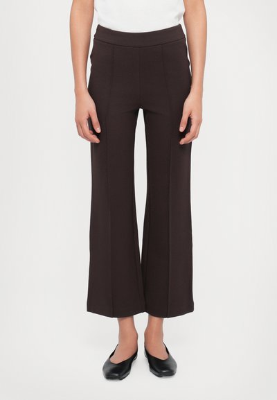 Pantalons marron à jambes larges en matériau extensible avec une taille haute et des coutures sur le devant. Associés à des ballerines noires.