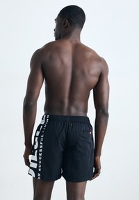 Black swim shorts featuring a side graphic print in white text, elastic waistband, and a back pocket with a flap and eyelet.