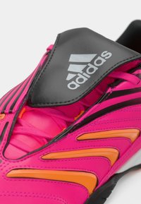 Κοντινό πλάνο ενός ροζ και πορτοκαλί ποδοσφαιρικού παπουτσιού Adidas με μαύρο καπάκι γλώσσας που φέρει το λογότυπο της Adidas σε ασημί.