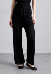 Pantaloni larghi in rete nera con vita alta e dettaglio a fiocco. Tessuto liscio con vestibilità ampia, abbinati a scarpe basse nere.