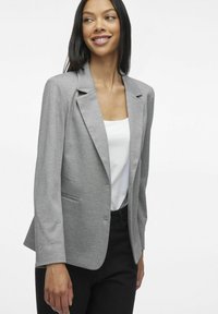 Blazer gris en tissu doux avec une coupe ajustée, fermeture à un bouton et deux poches avant, associé à un haut blanc.