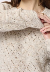 Gros plan sur une personne ajustant la manche d'un pull en tricot crème avec un motif en losange, portant une bague et un collier en or.