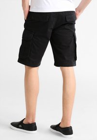 Shorts cargo en coton noir avec deux poches latérales et deux poches arrière, offrant une coupe décontractée et une longueur jusqu'aux genoux. Chaussures noires à enfiler.