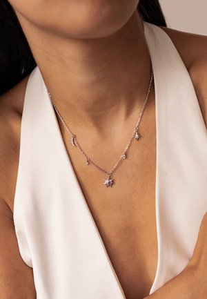 Silver necklace with celestial charms including a sun, star, crescent moon, and small crystals worn with a white halter top.