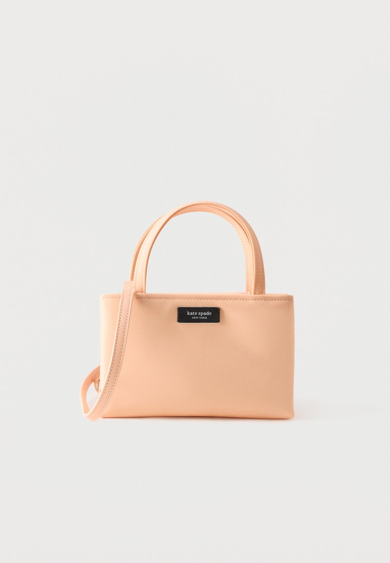 Μικρή ορθογώνια τσάντα ροδακινί με διπλές λαβές, αποσπώμενο ιμάντα ώμου και μαύρη ετικέτα "kate spade new york" στο μπροστινό μέρος.