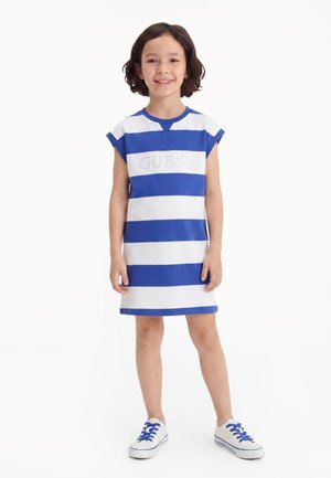 Dziecko stoi uśmiechnięte w niebiesko-białą pasiastą sukienkę bez rękawów i białe tenisówki z niebieskimi sznurowadłami na białym tle.
