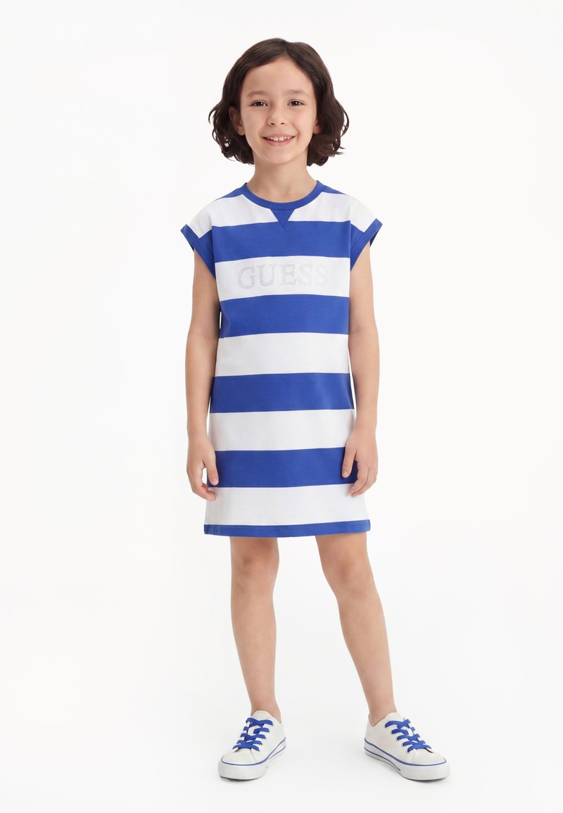 Ένα παιδί στέκεται χαμογελαστό ντυμένο με μπλε και λευκό ριγέ αμάνικο φόρεμα και λευκά αθλητικά παπούτσια με μπλε κορδόνια σε λευκό φόντο.