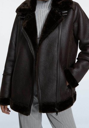 Femme portant une veste en peau retournée marron foncé sur un pull à col roulé côtelé gris et un pantalon gris à fines rayures, main dans la poche.