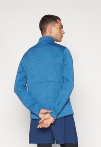 Pullover atletico blu con colletto alto, maniche lunghe e tessuto texturizzato. Presenta un gradiente di colore lungo la schiena e aperture per il pollice ai polsini.