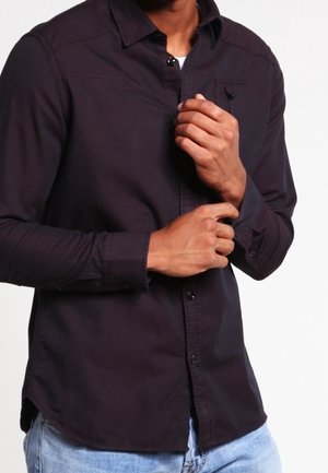 Mann trägt dunkles, langärmliges Hemd mit Knöpfen, richtet Manschette, kombiniert mit hellblauen Jeans, vor weißem Hintergrund.