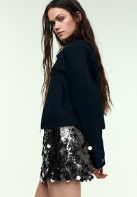 Marineblaue Cropped-Jacke mit strukturiertem Design, kombiniert mit einem schwarzen Minirock mit Pailletten in Form von Kreisen für eine strukturierte Optik.