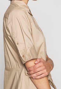 Beige kortärmad skjorta med knäppkrage och uppvikta ärmar; slät textur; med knytbar midja för justerbar passform.